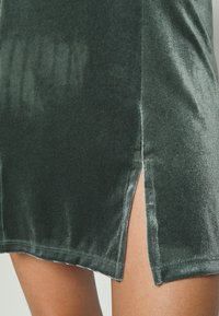 Tmavě zelené sametové šaty se spodním rozparkem, hladkým povrchem a délkou nad kolenem. Viditelné minimální švy.