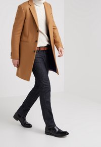 Man som går klädd i en kamelrock, krämfärgad kabelstickad polotröja, mörka jeans, brun bälte och svarta läder Chelsea boots på en vit bakgrund.