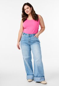 Pinkes Tanktop, hellblaue, weit geschnittene Jeans mit fransigen Säumen, kombiniert mit beigen Schuhen. Das Model hat wellige Haare und die Hände in den Jeanstaschen.