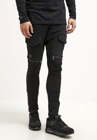 Svarta joggingsbyxor i stretchmaterial, med dragkedjefickor, ribbade knädetaljer och en smal passform. Bärs med svarta sneakers.