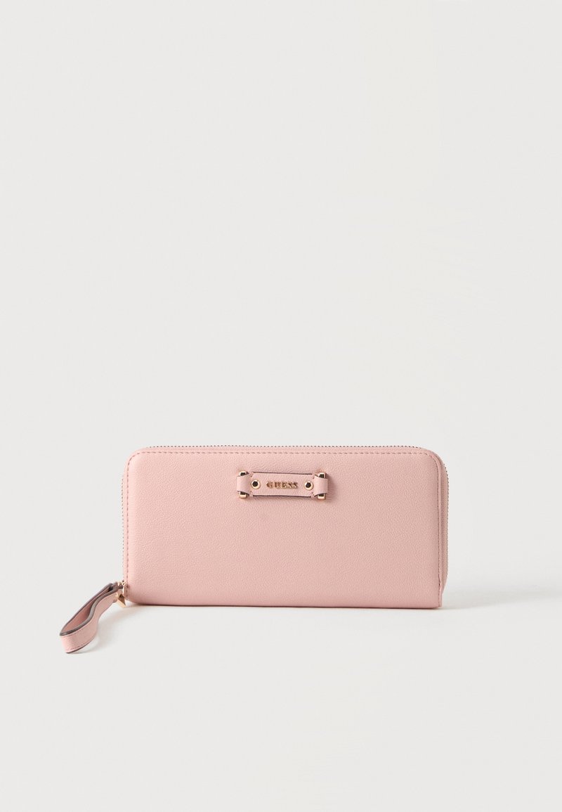 Portefeuille rose à fermeture éclair tout autour avec bracelet pour poignet et plaque métallique avec le logo de la marque sur le devant, sur un fond clair uni.