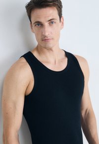 Top noir sans manches au design ajusté, fabriqué en tissu lisse. Doté de larges bretelles et d'un col rond. Le modèle a la peau claire et les cheveux courts.