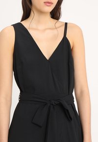 Musta hihaton haalari, jossa on V-aukko, istuva yläosa ja vyötäröön solmittava nauha. Sileä pinta ja virtaava siluetti.