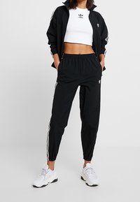 Svart träningsdress med vita detaljer, bestående av en kortärmad topp, zip-up jacka och slim-fit byxor med tre ränder på sidorna.