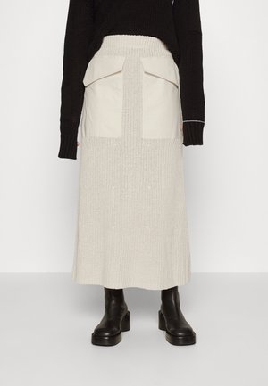Femme portant une jupe midi en maille crème avec de grandes poches avant, des bottes noires à talons épais et un pull noir à manches longues.