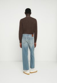 Καφέ ριμπ πλεκτό πουλόβερ, ανοιχτό μπλε τζιν παντελόνι με φαρδιά πόδια και δερμάτινη ταμπέλα στο πίσω μέρος. Λευκά αθλητικά παπούτσια με μπεζ σόλα, με φόντο απλό.