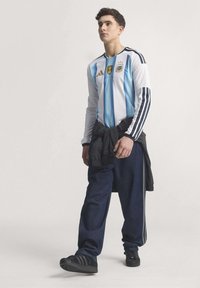 Bijela majica s svijetloplavim vertikalnim prugama, s Adidas logotipom i grbom tima, u kombinaciji s tamnim trapericama i crnim cipelama.