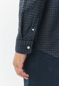 Camicia a quadri nera e grigia con polsino abbottonato, tessuto strutturato e design aderente. Pantaloni blu parzialmente visibili.
