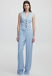 Gilet azzurro su misura con scollo a V e cinque bottoni scuri, abbinato a pantaloni a vita alta e gamba larga dello stesso colore. Tessuto liscio.
