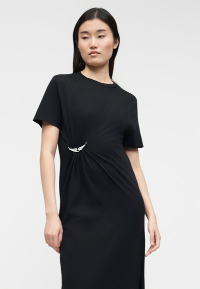 Γυναίκα που φοράει μαύρο φόρεμα με κοντά μανίκια, με ασημένιο κούμπωμα σε σχήμα φτερού που συγκεντρώνει το ύφασμα κάτω από το στήθος, πάνω σε απλό γκρι φόντο.