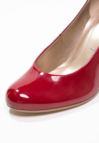 Chaussure à talon haut rouge brillant avec bout arrondi et intérieur beige, vue en gros plan sous un angle.