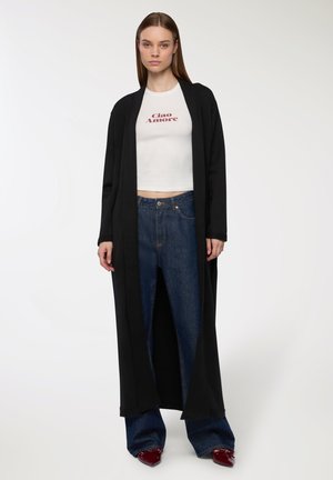 Lungo cappotto nero sopra una maglietta bianca corta con il testo "Ciao Amore", abbinato a jeans blu a vita alta e scarpe rosse a punta.