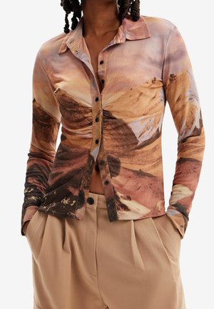 Woman wearing a ruched button-up shirt with earthy abstract print and beige high-waist pleated trousers, hands in pockets.