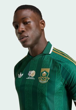 Jonge man die een groen voetbalshirt van Zuid-Afrika draagt met gouden logo's en bies tegen een lichte achtergrond.