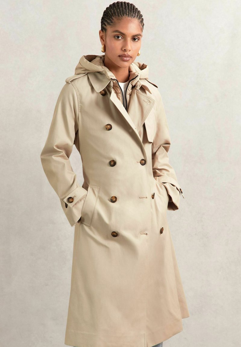 Beige trenchcoat med huva, dubbelsidig design, stora bruna knappar och sidofickor. Robust tyg med en slät textur.