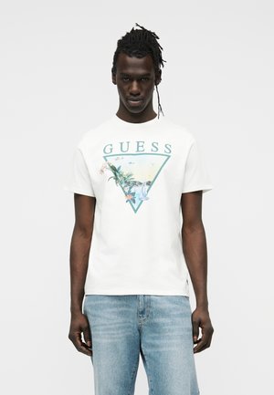 Homme aux dreadlocks portant un t-shirt blanc Guess avec graphisme floral et de voilier, associé à un jean bleu clair, se tenant devant un fond uni.