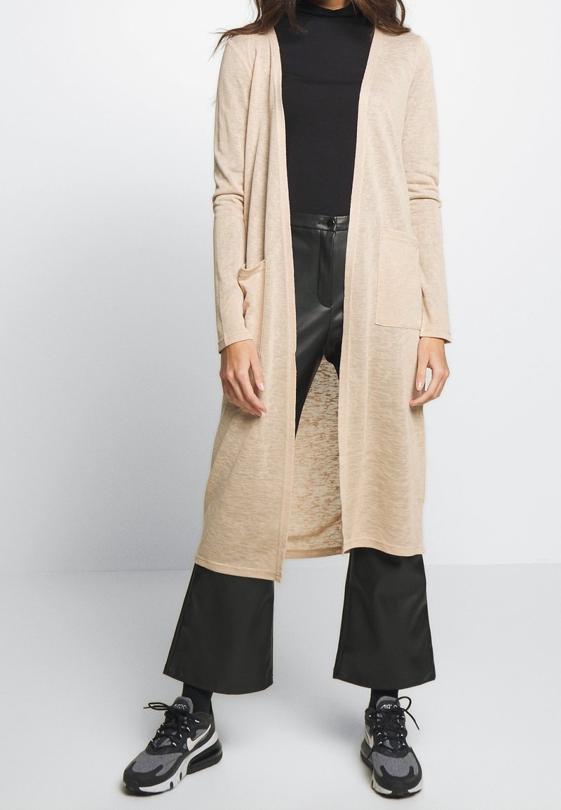 Personne portant un long cardigan beige avec poches, un haut noir à col montant, un pantalon noir à jambes larges, et des baskets noires et grises.