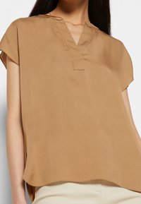 Beige Bluse mit kurzen Ärmeln aus einem leichten, glatten Stoff, mit V-Ausschnitt und seitlichen Schlitzen. Einfaches Design mit minimalen Details.