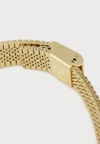 Montre-bracelet en métal doré avec un bracelet en maille tressée, fermoir lisse avec texte gravé, et un détail de design en relief sur les bords du bracelet.