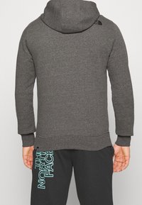 Felpa grigia con cappuccio, maniche lunghe, orlo a coste e dettaglio del logo sulla spalla. Abbinata a pantaloni neri con design di testo azzurro chiaro.