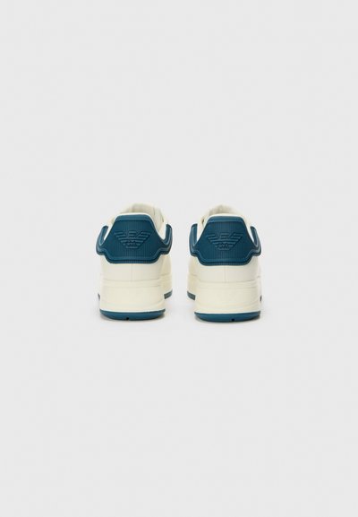 Baskets blanches avec un accent de talon bleu texturé, présentant un dos avec une marque, un dessus lisse semblable à du cuir et une semelle en caoutchouc bleue.