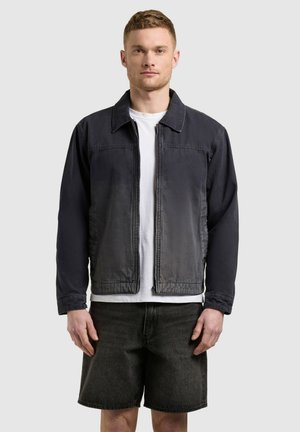 Muž v čiernej bunde na zips cez biele tričko a čierne džínsové šortky, stojaci pred jednoduchým svetlým pozadím.