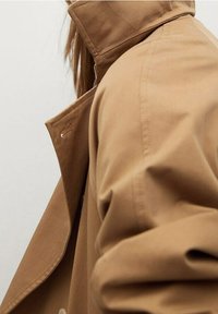 Trench-coat beige à col haut, avec une texture lisse et des détails de coutures visibles. Vue de côté mettant en valeur le tissu drapé et les contours.