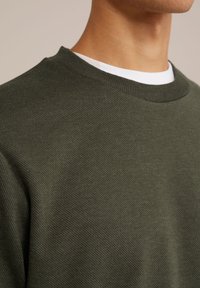 Sudadera verde oliva con un patrón texturizado y acanalado, que presenta un cuello redondo y una capa interior blanca visible en el borde del cuello.