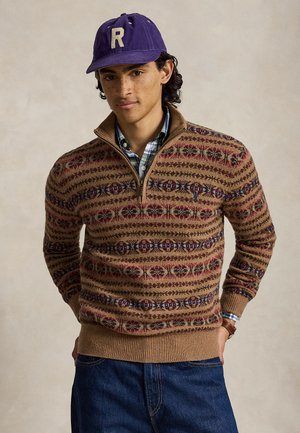 Jeune homme aux cheveux bouclés porte une casquette violette avec la lettre R, un pull marron à fermeture éclair partielle à motifs par-dessus une chemise à carreaux et un jean bleu.