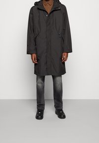 Impermeable gris con capucha, bolsillos con cremallera y cierre a presión. Lleva encima una camisa marrón y jeans grises, combinados con zapatos negros.