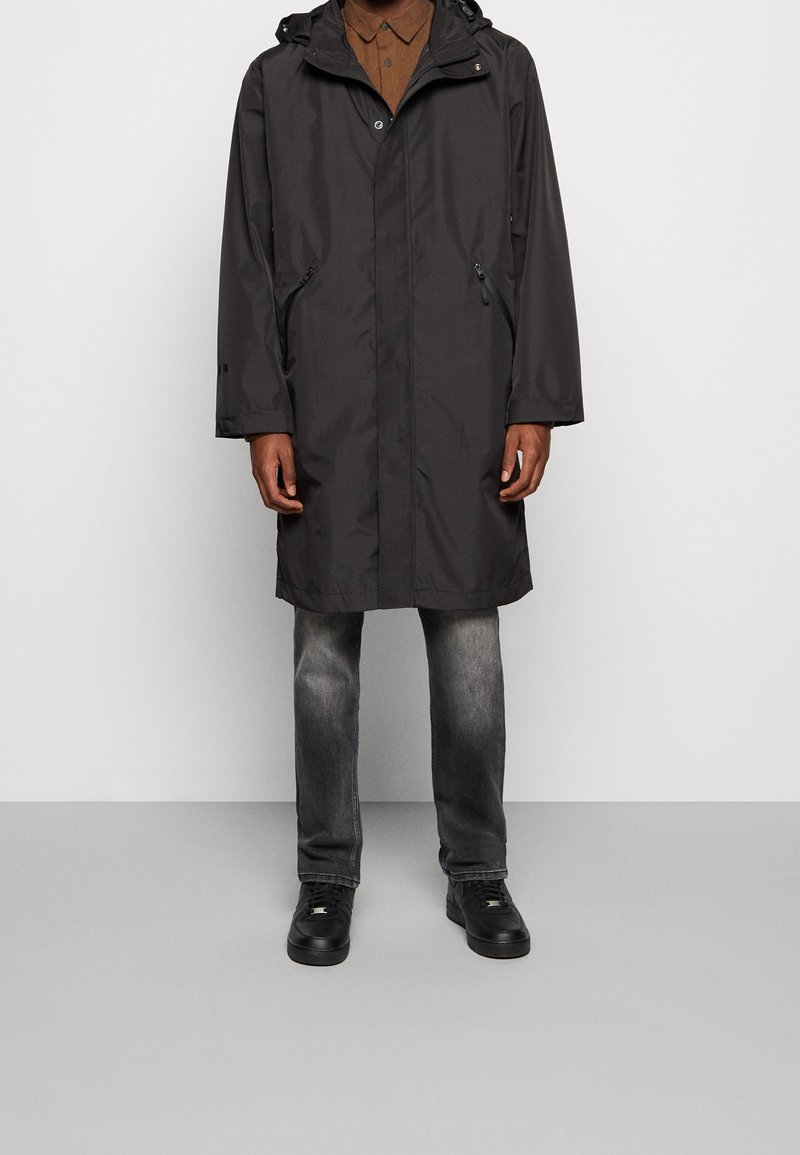 Impermeable gris con capucha, bolsillos con cremallera y cierre a presión. Lleva encima una camisa marrón y jeans grises, combinados con zapatos negros.