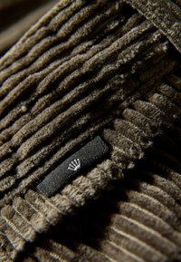 Bruine corduroy-stof met opvallende verticale richels, met een klein zwart label met een zilveren embleem in de linkerbenedenhoek.