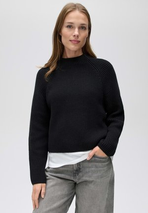 Vrouw met lichtbruin haar, die een zwarte gebreide trui draagt over een witte blouse en lichtgrijze spijkerbroek, staand tegen een eenvoudige lichte achtergrond.