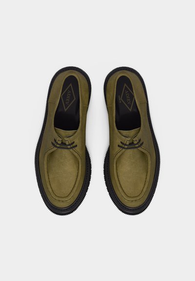 Ζεύγος μοκasinιών από ελιά σουέτ με μαύρες ελαστικές σόλες και μαύρα κορδόνια, σε θέα από πάνω σε λευκό φόντο.