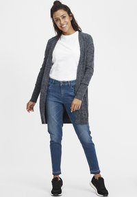 Garš, atvērtā priekšpuse pelēkā tumši dzijas kardigans, kombinēts ar baltu T-kreklu un ziliem pieguļošiem džinsiem. Valkāts ar melnām sporta kurpēm.