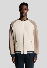 Beige lynlåsjakke med raglanærmer i lys beige og marineblå. Har sidelommer, ribbede manchetter og et diskret logo på brystet.