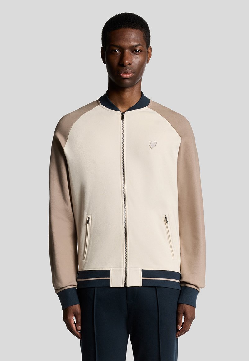 Beige lynlåsjakke med raglanærmer i lys beige og marineblå. Har sidelommer, ribbede manchetter og et diskret logo på brystet.