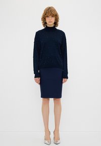 Marine-blauer Strickrollkragenpullover verziert mit kleinen strukturierten Perlen, kombiniert mit einem figurbetonten marineblauen Rock und silbernen spitzen Absatzschuhen.