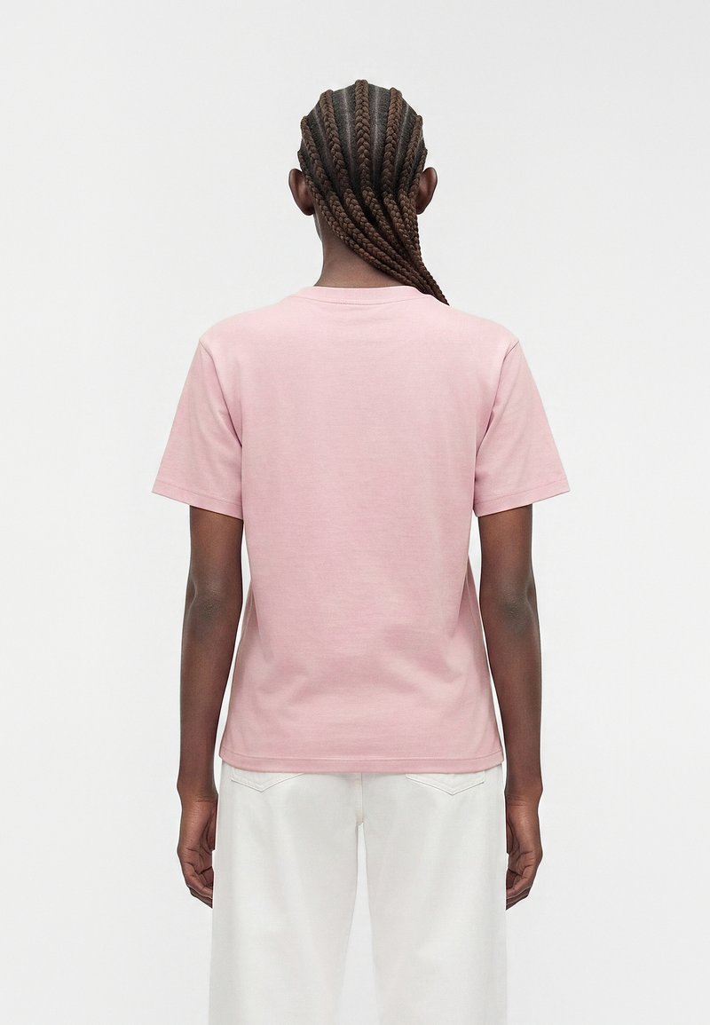 Person with long braids wearing a light pink short-sleeve shirt and white pants, standing facing away against a plain background.