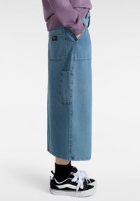 Falda de mezclilla azul clara con bolsillos laterales, a la altura de la rodilla y de corte relajado. Combinada con zapatillas negras y blancas y calcetines negros de caña corta.