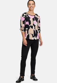 Langärmlige Bluse mit abstraktem Druck in Creme, Schwarz und Pink. Weicher Stoff, lockerer Schnitt und runder Ausschnitt. Kombiniert mit schwarzen Hosen und Schuhen.