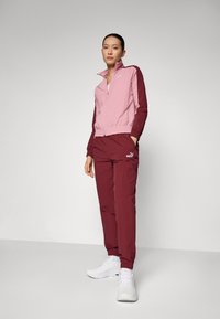 Puma sportska odjeća u ružičastoj i tamnocrvenoj boji. Jakna sa zatvaračem ima elastične manžete i pojas. Hlače također imaju elastični pojas i detalj s logotipom. Bijele tenisice.