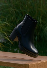 Svart läderankelboots med klumpig klack, rundad tå, vertikal söm och en detalj med sidodragkedja. Mot en naturlig bakgrund.