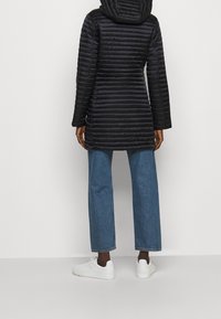 Schwarze Puffjacke mit vertikalen Nähten, einer Kapuze und langen Ärmeln, kombiniert mit blauen weiten Jeans und weißen Sneakers.