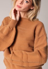 Femme blonde avec un maquillage léger portant un pull oversize texturé de couleur camel, touchant son cou de sa main gauche.