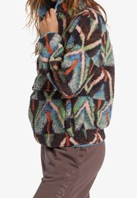 Pehmeä, monivärinen fleece-takki, jossa on abstrakteja kuvioita vihreänä, vaaleanpunaisena, ruskeana ja sinisenä. Vetoketjullinen etuosa ja korkea kaulus.