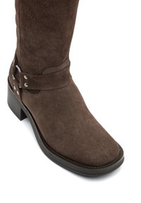 Bota de ante marrón hasta la rodilla con punta cuadrada, que presenta una correa decorativa y detalles en plata, además de un tacón grueso. Superficie texturizada.