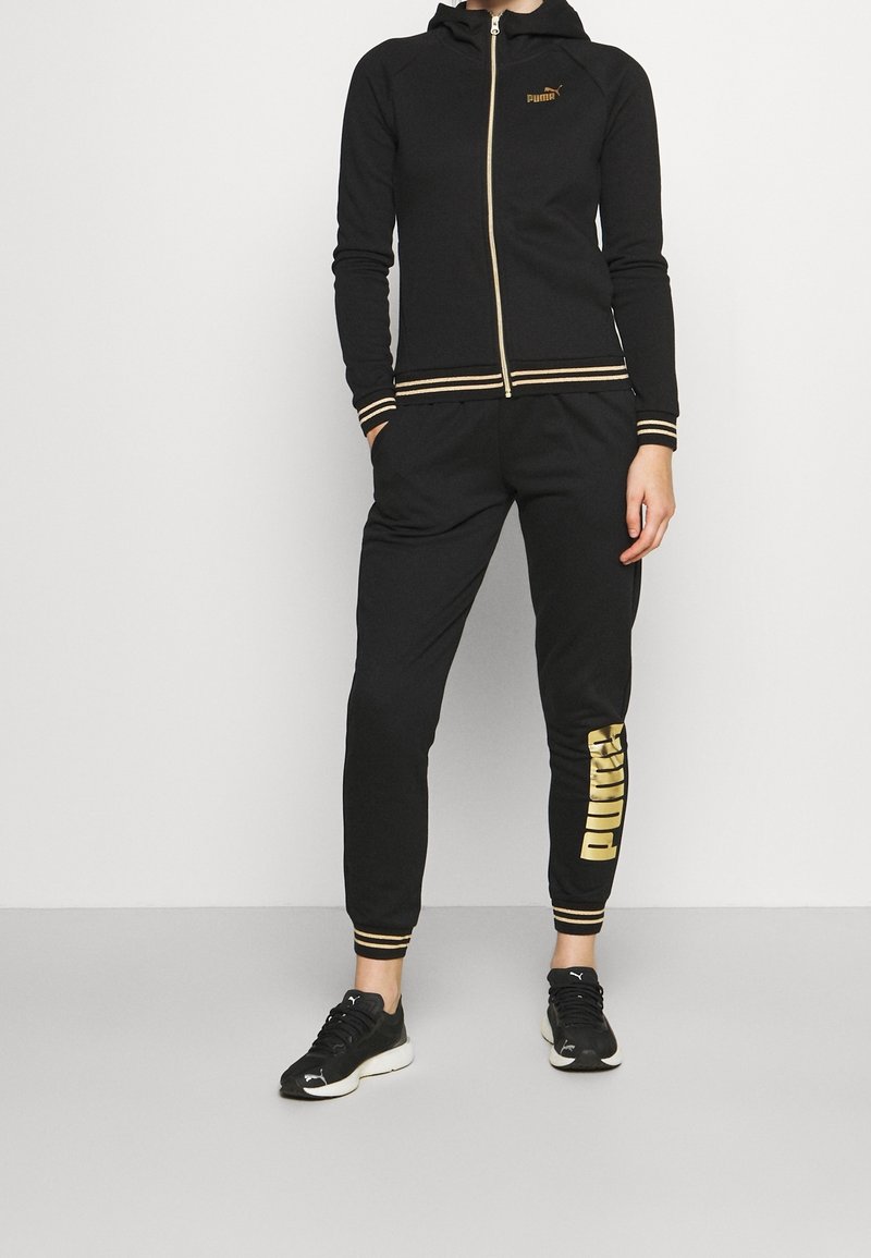 Personne portant un survêtement Puma noir avec des accents dorés, comprenant un sweat à capuche zippé et un jogging avec "PUMA" en or sur une jambe.