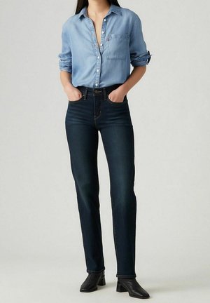 Kvinde iført en lyseblå skjorte med knapper stukket ned i mørkeblå højtaljede jeans med sorte ankelstøvler med hæl, hænderne i lommerne.