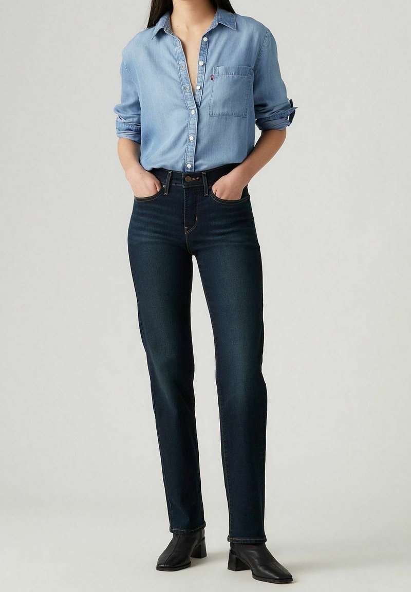 Femme portant une chemise bleu clair boutonnée rentrée dans un jean taille haute bleu foncé, avec des bottines noires à talons, les mains dans les poches.